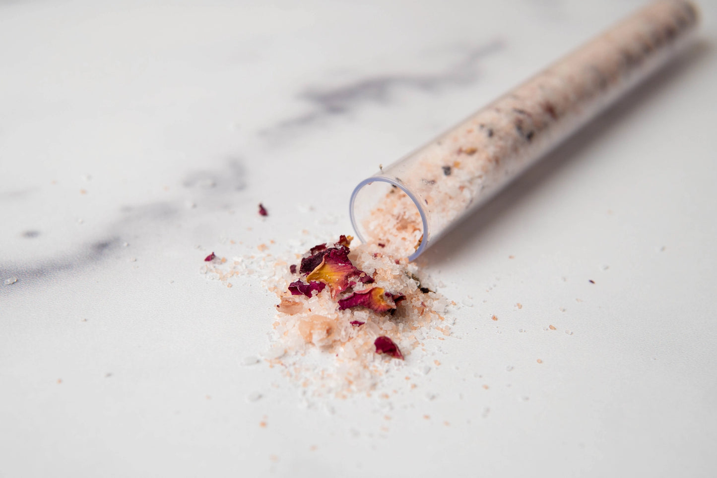 Bath salt with rose petals spilling out of a glass container on a marble surface